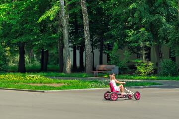 Little girl riding pink pedal car in sunny summer park.Happy childhood: A young girl enjoys ride on her go-kart outdoors.Active kid playing in the park, driving a four-wheeled toy car on a summer day