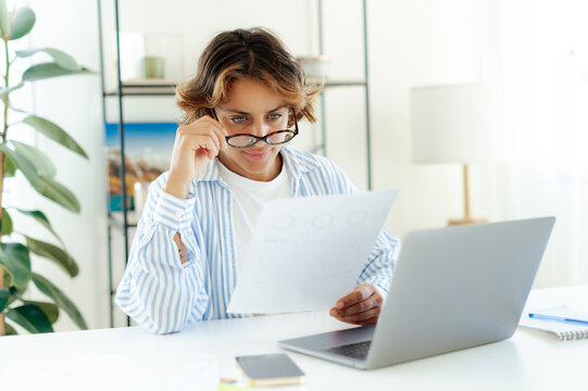 Serious brazilian successful businesswoman with glassed reading printed report while working on laptop at home. Focused professional analyzing data or financial charts during remote work in apartment