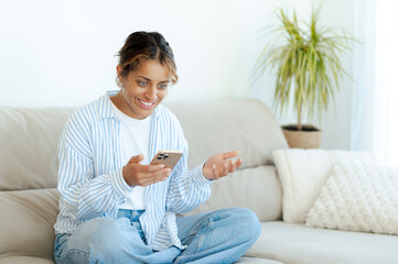 Excited happy brazilian woman sits cross-legged on sofa, laughing and chatting during video call on...