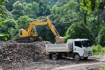 A backhoe is scooping up a large pile of rocks onto a white truck to build a road barrier.