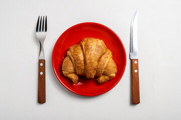 Spoon and fork with freshly baked croissant on a red plate with a white background. Top view