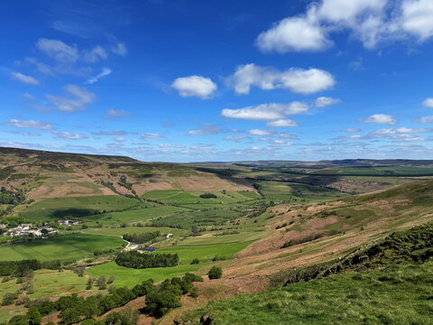 Panoramic view from Mam Tor ridge of green valleys and rolling hills of the Peak District under a bright blue sky with clouds. England, UK.