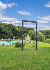 Girl in a hoodie relaxing on a large modern swing at a summer resort overlooking a pool and green forest.Summer leisure and recreation concept. A person rests on a swing in a beautiful resort area