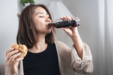 Asian woman drinking soda and holding a burger in both hands, showing emotional eating and unhealthy food choices high in sugar, salt, and fat