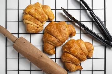 Tongs and rolling pin with freshly baked croissants on a cooling tray with a white background. Top...