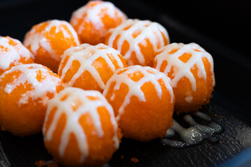 Sushi balls covered in bright orange tobiko and drizzled with white mayonnaise on a black tray, highlighting texture and vibrant color.