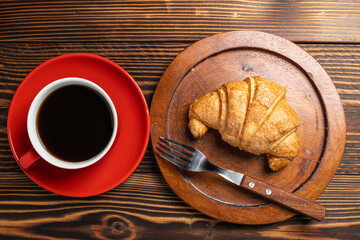 Fork and a freshly baked croissant on a wooden tray with a cup of coffee on a wooden background. Top view