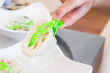 Hand holding sliced lotus root with green tongs over a white bowl filled with ice, showing fresh preparation of an Asian vegetable.