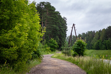 A narrow gravel path curves through a lush green forest with wooden power poles under a cloudy,...
