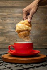 Closeup view human hand dipping a freshly baked croissant into a cup of coffee on a wooden tray...