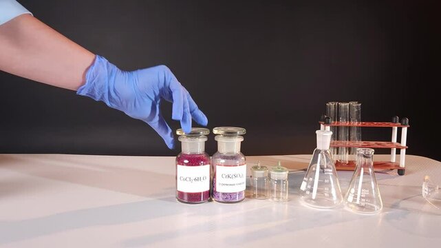 A girl's hand in a blue rubber glove places glass jars with multi-colored chemical compounds in powder form on the table
