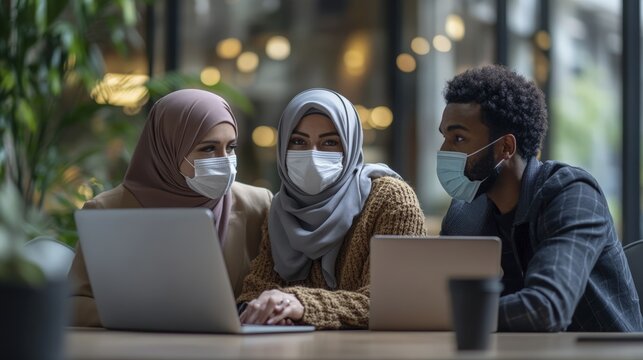 Diverse group wearing masks and hijabs collaborating on laptops in an office setting