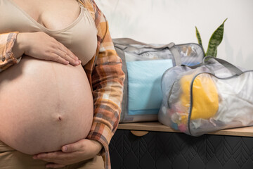 Pregnant woman stroke big belly and collects a bag in the hospital at home. Future mom at 9 months of pregnancy preparing and planning newborn baby clothes. Anticipation for the birth of a child