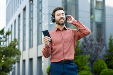 A man wearing headphones listens to music on his phone outside a modern building, enjoying the...