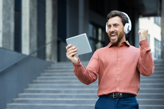 A happy man with a beard enjoys something on his tablet while wearing headphones outside. - Powered by Adobe