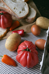 A rustic flat lay featuring slices of homemade artisan bread, fresh brown eggs in a carton, heirloom tomato, sweet peppers, and potatoes on a wooden kitchen table. Natural light and cozy.