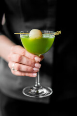 Elegant hand holding a green cocktail in a clear glass, garnished with lychee on a skewer, against...