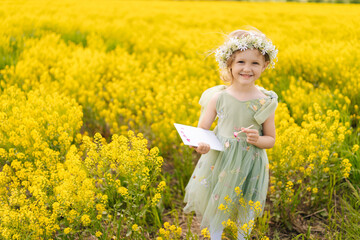 Cheerful child girl wearing flower wreath and fairy costume standing posing at blooming field...
