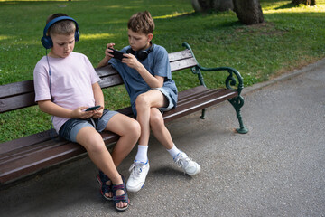 Two boys playing online games on smartphones with headphones in park sitting on bench, focused on...