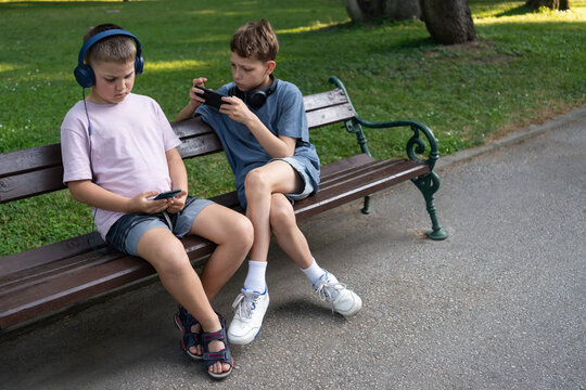 Two boys playing online games on smartphones with headphones in park sitting on bench, focused on phone screens. Digital lifestyle habits of modern children, virtual kids play, childhood tech culture
