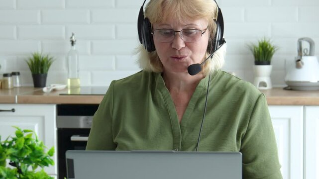 Focused employee wearing headphones with headset, speaking during virtual conference with laptop in kitchen, attending online meeting, participating in video call, looking at computer screen.