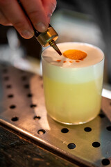 Close-up of a bartender adding bitters to a craft cocktail with orange garnish and frothy top,...