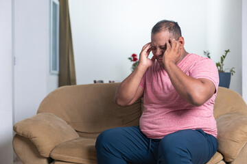A man in a red shirt is sitting on a couch and rubbing his head