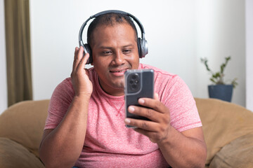 A man wearing a pink shirt is sitting on a couch and looking at his cell phone