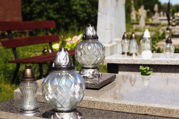 Grave with decorative glass lanterns. Cemetery memorial with candles on tombstone. Peaceful resting place. Daylight scene with greenery and bench. Symbol of memory and remembrance. Polish tradition.