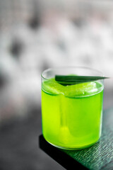 Refreshing green cocktail with ice cubes and a leaf garnish in a clear glass on a dark wooden surface, captured in a close-up with soft blur in the background