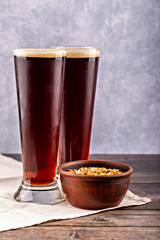 Two tall glasses of dark beer with creamy foam next to rustic clay bowl of salted peanuts. Wooden...