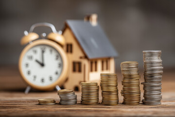 Stack of coins growing in size next to a wooden house model and a clock, symbolizing long-term savings and retirement planning, with copy space for economic concept