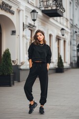 A stylish young woman in a black tracksuit sports suit mockup stands confidently on a city street. She has long hair and wears sneakers, with a modern urban backdrop featuring classic architecture.