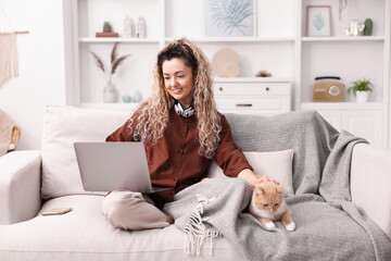 Obraz premium Smiling woman working with laptop and stroking cat on sofa in home office