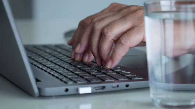 Close up hands quickly typing on laptop keyboard, with glass water placed conveniently next to computer on clean white desk, suggesting busy workday. Programmers job. Fingers typing prompt on keyboard