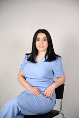 A beautiful, smiling female nurse in a blue medical suit sits on a chair on a white background. Portrait of a doctor. The image conveys professionalism and experience in the field of healthcare