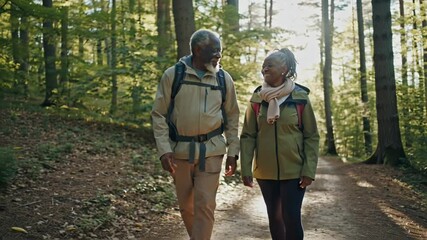 Happy black senior couple wearing backpacks walking along forest path, enjoying hike together in nature - Powered by Adobe