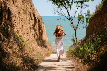 Woman in white dress and hat walks down a coastal path between cliffs toward the sea, enjoying a...