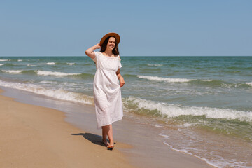 Smiling woman in white dress and hat walks barefoot along the beach shoreline, enjoying a sunny...