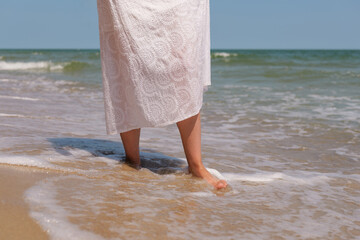 Close-up of a woman’s bare feet and white dress as she walks along the shoreline, feeling the...