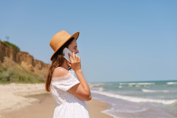 Young woman in a summer dress and hat talking on her smartphone while walking along a sunny beach...