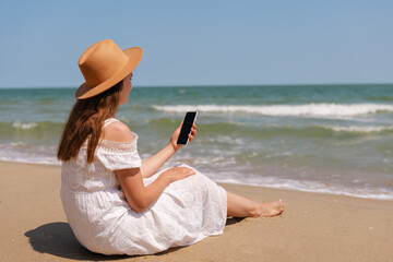 Woman in a white dress and hat sitting on the beach, holding a smartphone and looking at the sea on...