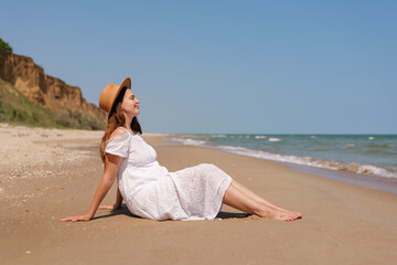 Young woman in white dress and hat relaxing on a sandy beach, enjoying the sun and sea breeze with...