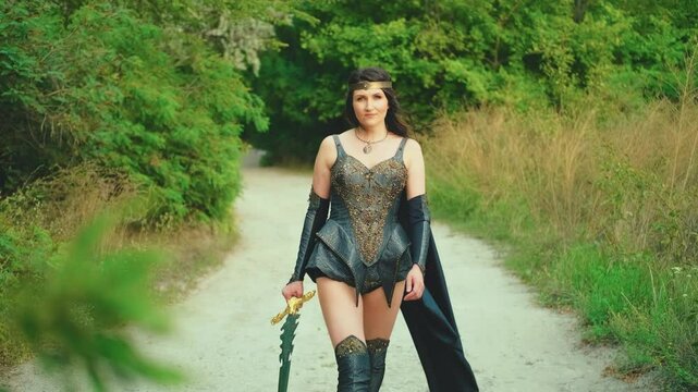 Portrait Fantasy woman viking princess warrior walks down road. girl old style sexy valkyrie holding sword in hand. Black Leather costume armor cloak flies in wind. green nature summer. art video 4k