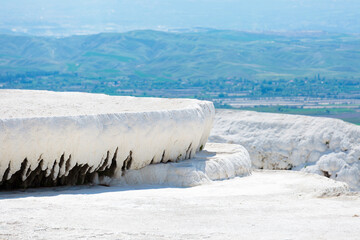 Natural travertine formation with white calcium deposits in Pamukkale, Turkey. Elevated terrace against scenic background of distant green hills under bright daylight