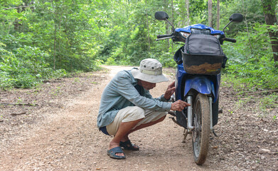 An elderly Asian man inspects a broken motorcycle on a dirt road in the forest.