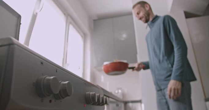 A young man flips food in a pan in his apartment kitchen. He is making breakfast, lunch, or dinner.