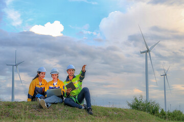 Team male and female engineers sit together grassy hill at wind farm sharing concept about...