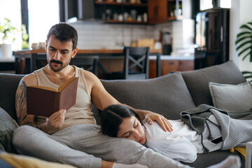 Young couple relaxing on the sofa at home, man reading a book while his girlfriend is sleeping with her head on his legs