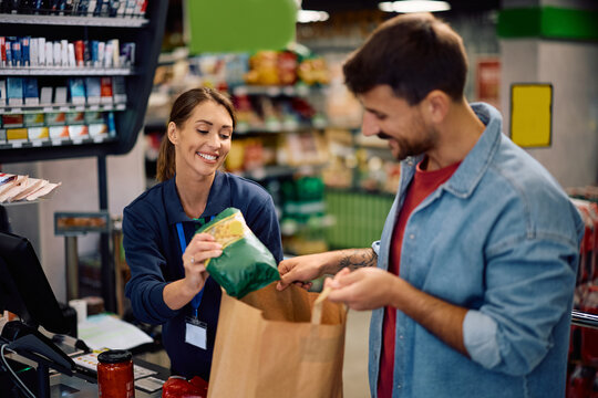 Happy cashier helping a customer with packing goods in paper bag at supermarket checkout. - Powered by Adobe